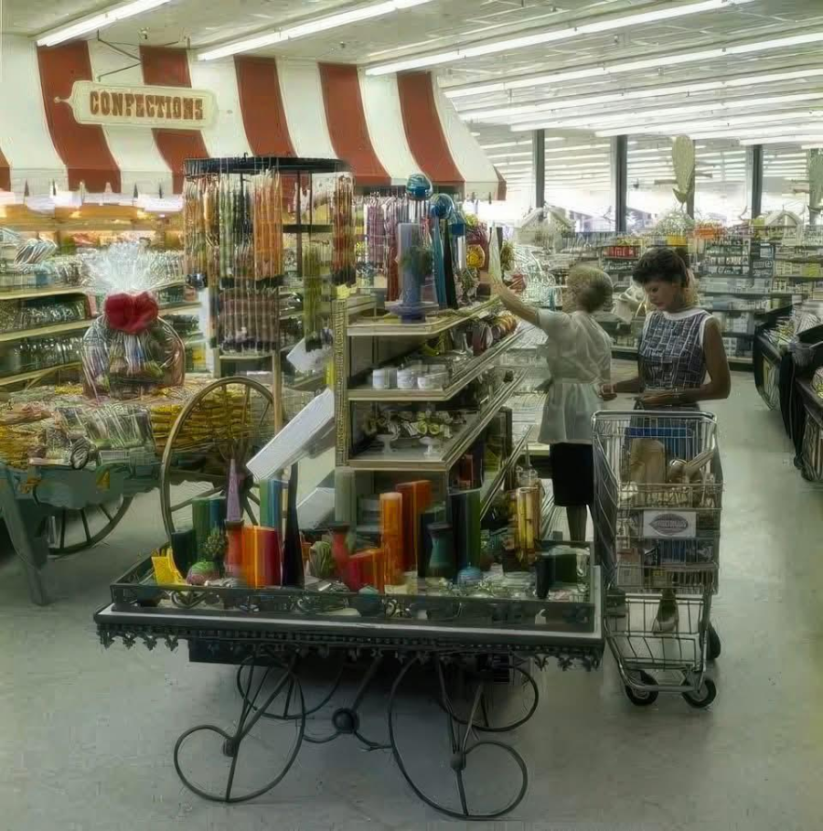 Two women with a shopping cart browse colorful candles and goods in a retro grocery store. The store has bright lighting, striped decor, and a sign reading "CONFECTIONS" above candy displays.