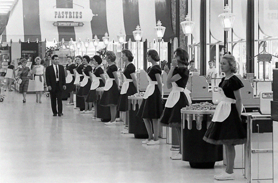 Black and white photo of women in matching uniforms and aprons standing at cash registers in a row inside a large, brightly lit supermarket, with a man in a suit walking between them.
