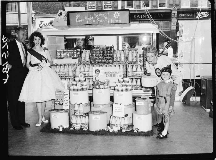 A black-and-white photo shows people posing by a supermarket dairy display with milk bottles, cheese, and butter. A woman in a sash, a man in a suit, another woman, and a young girl stand smiling in front of the products.