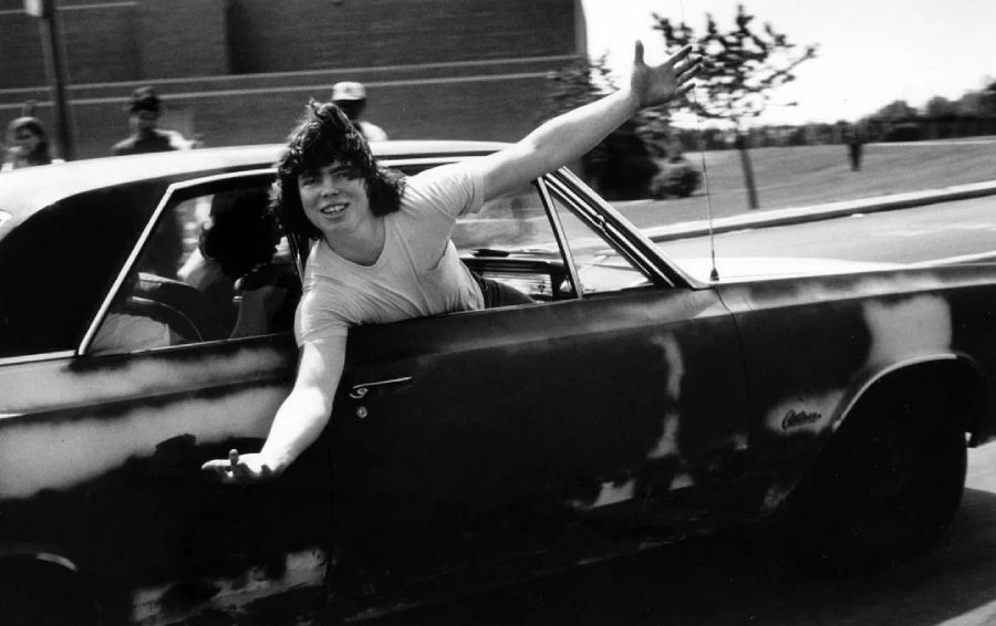 A young man leans out of the driver's window of an old car, smiling with his arms outstretched. The car's paint is worn and patchy. Trees, a building, and people are visible in the background on a sunny day.