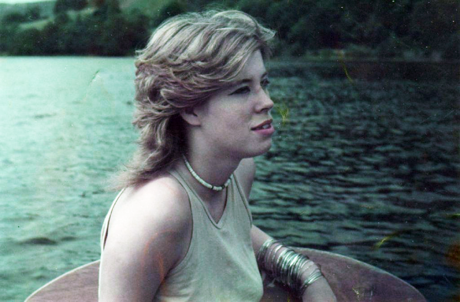 A young woman with short, wavy, light-colored hair sits on a wooden boat, wearing a light tank top, necklaces, and bangles, gazing thoughtfully over a calm lake with trees in the background.