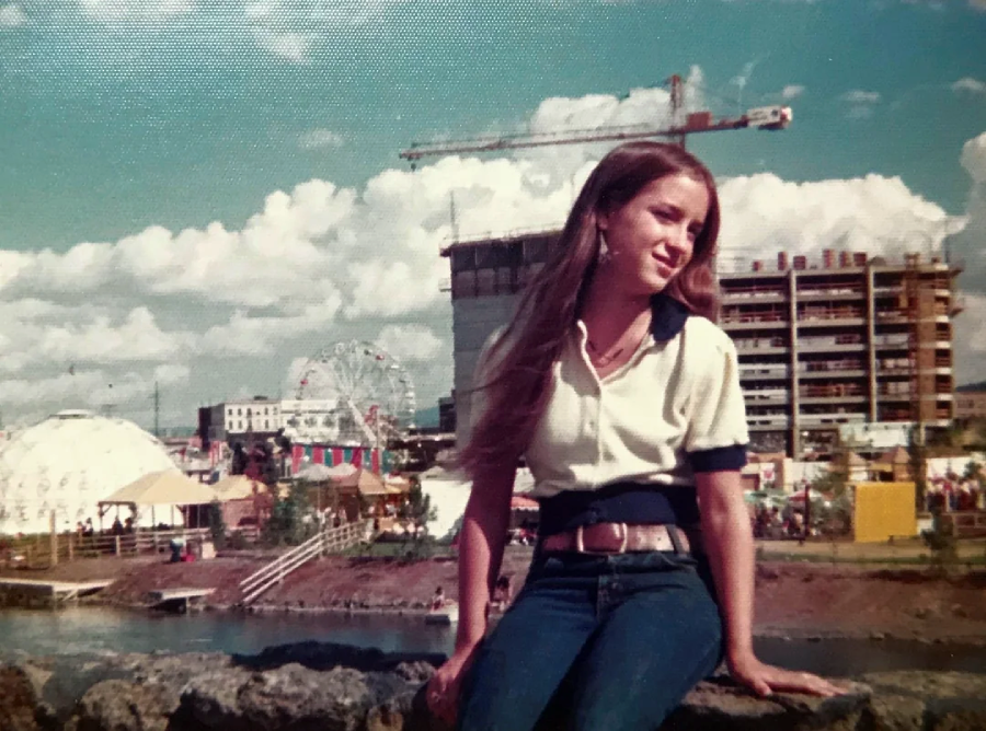 A young woman with long hair sits on a stone wall, smiling, with a construction site, ferris wheel, and fair tents visible in the background under a partly cloudy sky.