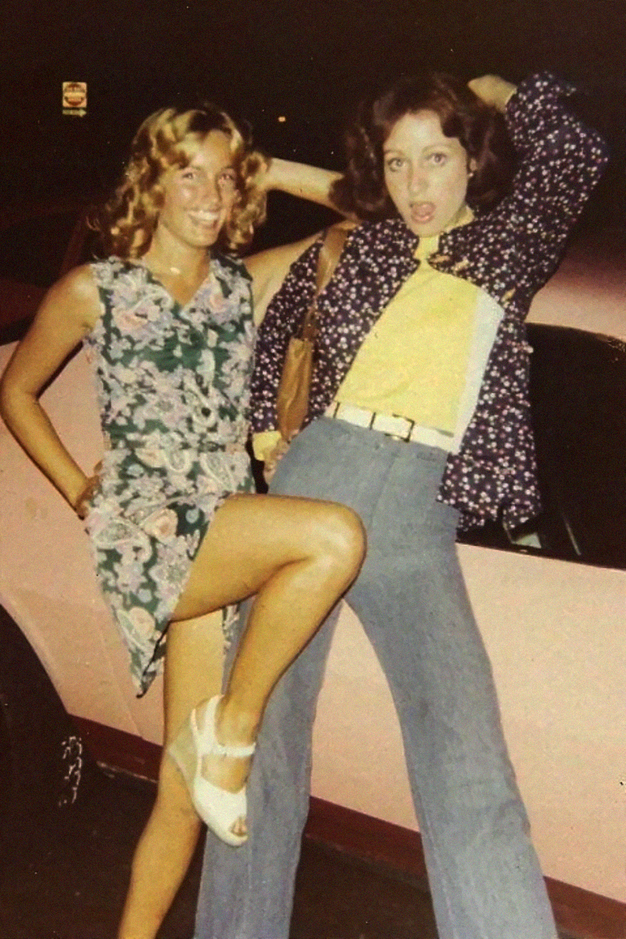 Two young women pose playfully in front of a car at night. One wears a floral dress and white heels, lifting her leg. The other wears flared jeans, a yellow top, and a patterned jacket, striking a dramatic pose.