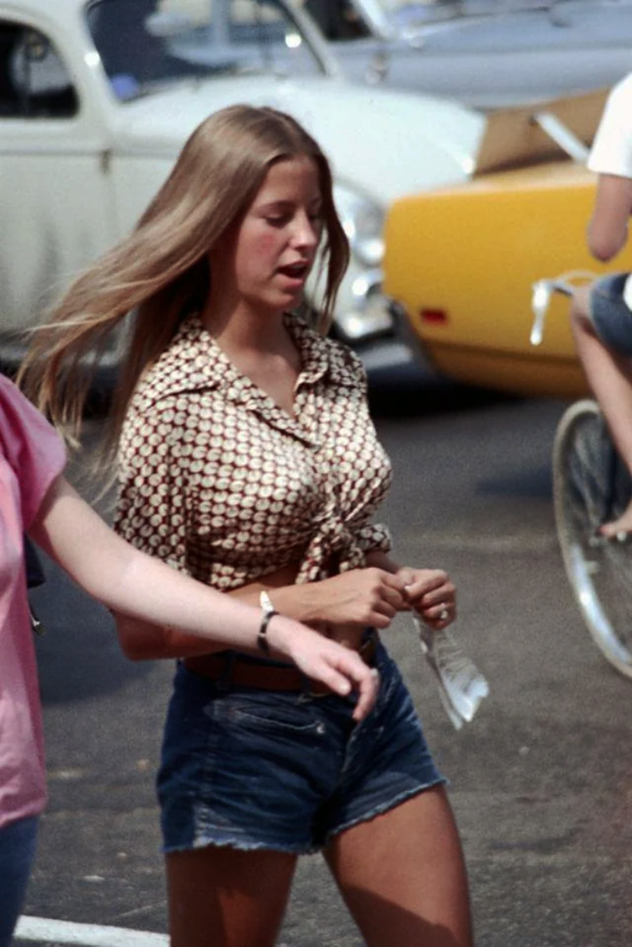 A young woman with long hair wears a patterned, knotted shirt and denim shorts while walking outdoors on a sunny day. Vintage cars and a person on a bicycle are visible in the background.