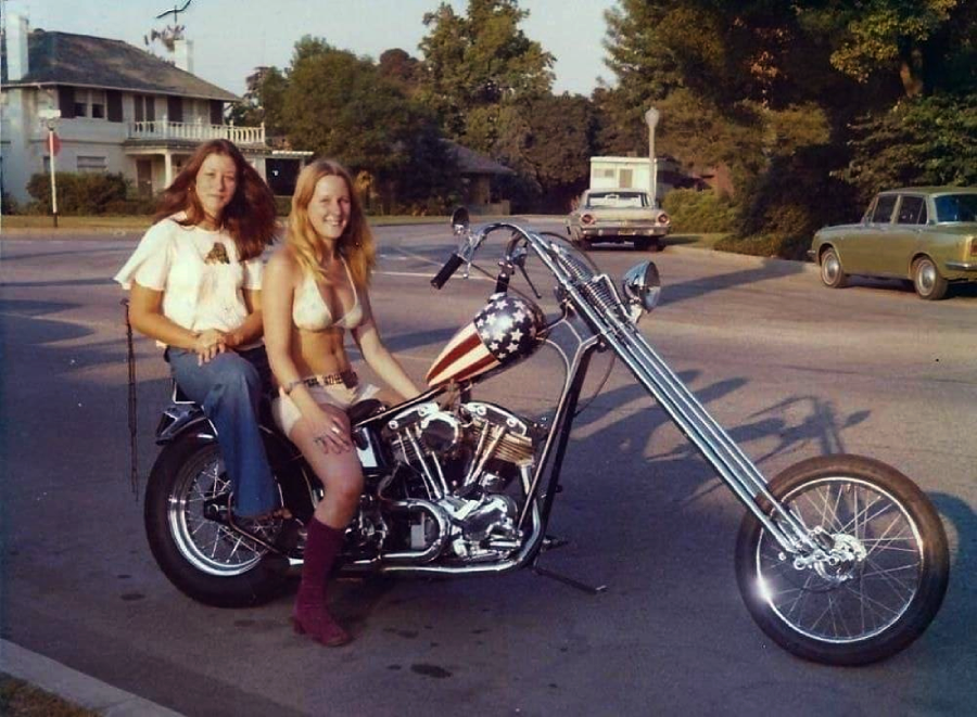 Two women sit on a custom chopper motorcycle with an American flag design on the gas tank, parked on a street in a suburban neighborhood with vintage cars and trees in the background.