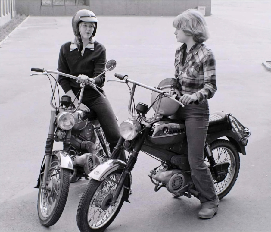 Two young women sit on vintage mopeds, facing each other and talking. One wears a helmet and sweater, the other holds a helmet and wears a plaid shirt. They are in an open paved area. The photo is black and white.