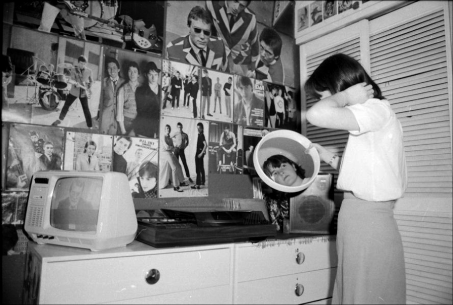 A person stands at a dresser, looking into a small round mirror. The wall behind is covered with posters of musicians and bands. A TV sits on the dresser, displaying a black-and-white image.
