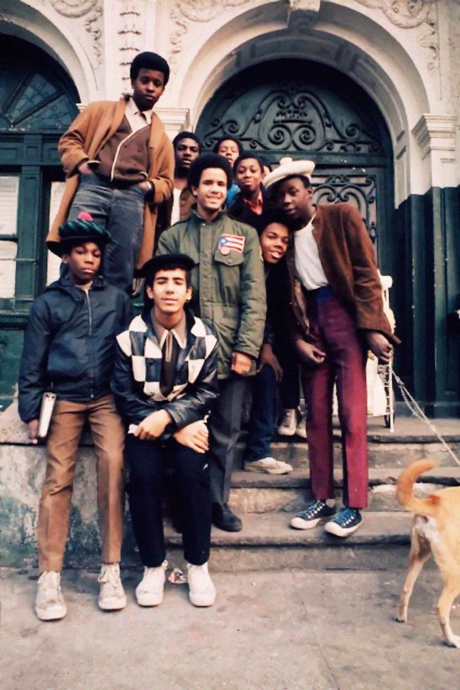 A group of young boys pose together on the steps of a building with ornate doors. Some stand, some crouch, and one holds a dog on a leash. They wear a mix of jackets, hats, and casual clothes.