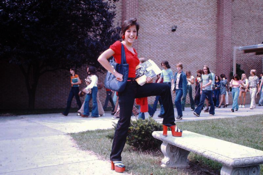 A smiling woman in a red top and black pants poses with one foot on a bench, showing off red platform shoes. She holds books and a bag, with a group of casually dressed people walking behind her outside a brick building.