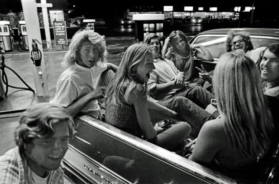 A group of young people laugh and talk while sitting in the back of a convertible car at a gas station at night. The scene is lively, with gas pumps and a lit-up building visible in the background.