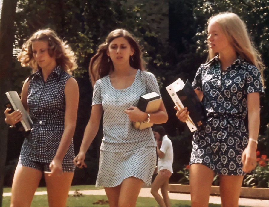 Three young women in patterned dresses walk outside on a sunny day, each holding books and folders. Trees and greenery are visible in the background, with another person seated on grass behind them.