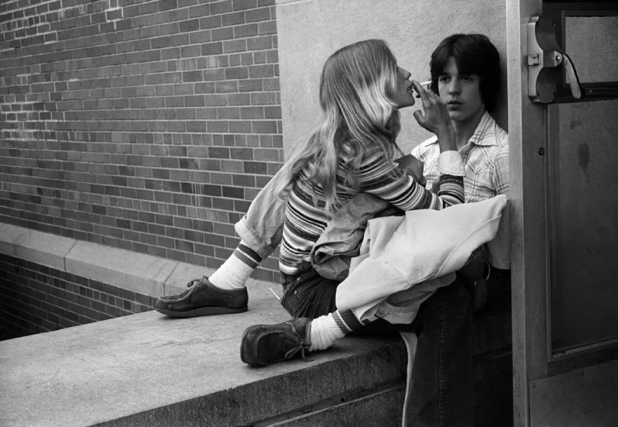 A teenage girl sits close to a teenage boy on a concrete ledge by a brick wall. She gently touches his chin, while he looks forward with a neutral expression. Both wear casual clothes and appear to be in a candid moment.