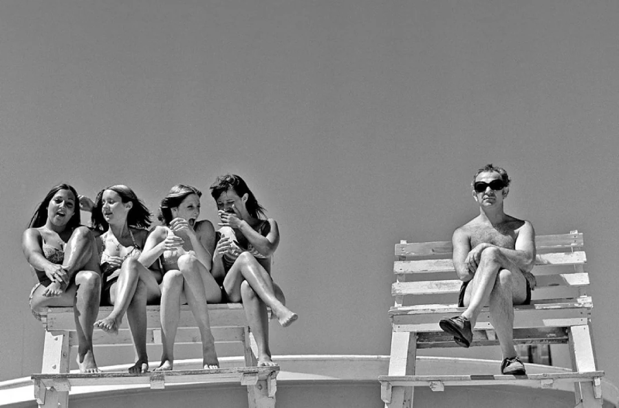 Four women in swimsuits sit together on a bench, talking and laughing, while a man wearing sunglasses sits alone on a separate bench, looking away with crossed legs. The sky is clear in the background.