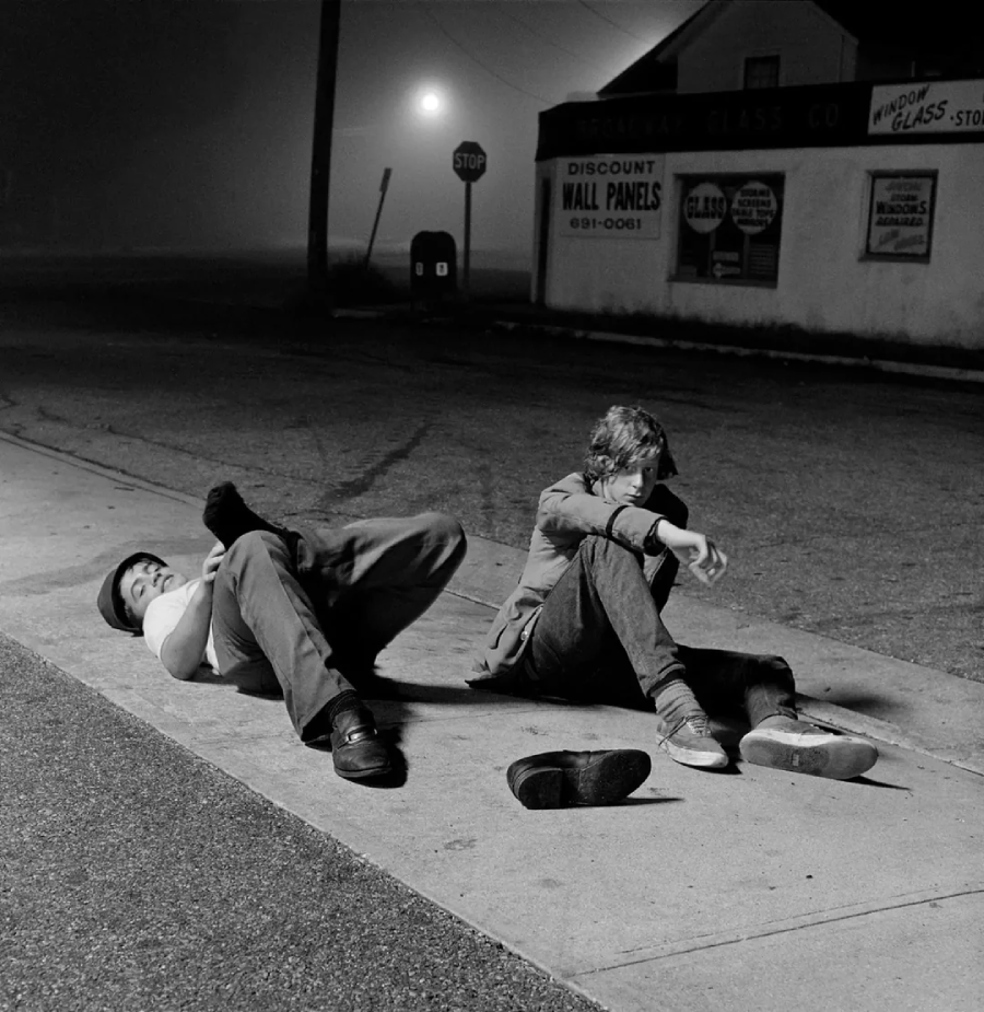 Two people sit and lie on a sidewalk at night under a streetlight. One person's shoes are off and lying nearby. The street is empty, and buildings with signs are visible in the background. The scene appears quiet and deserted.