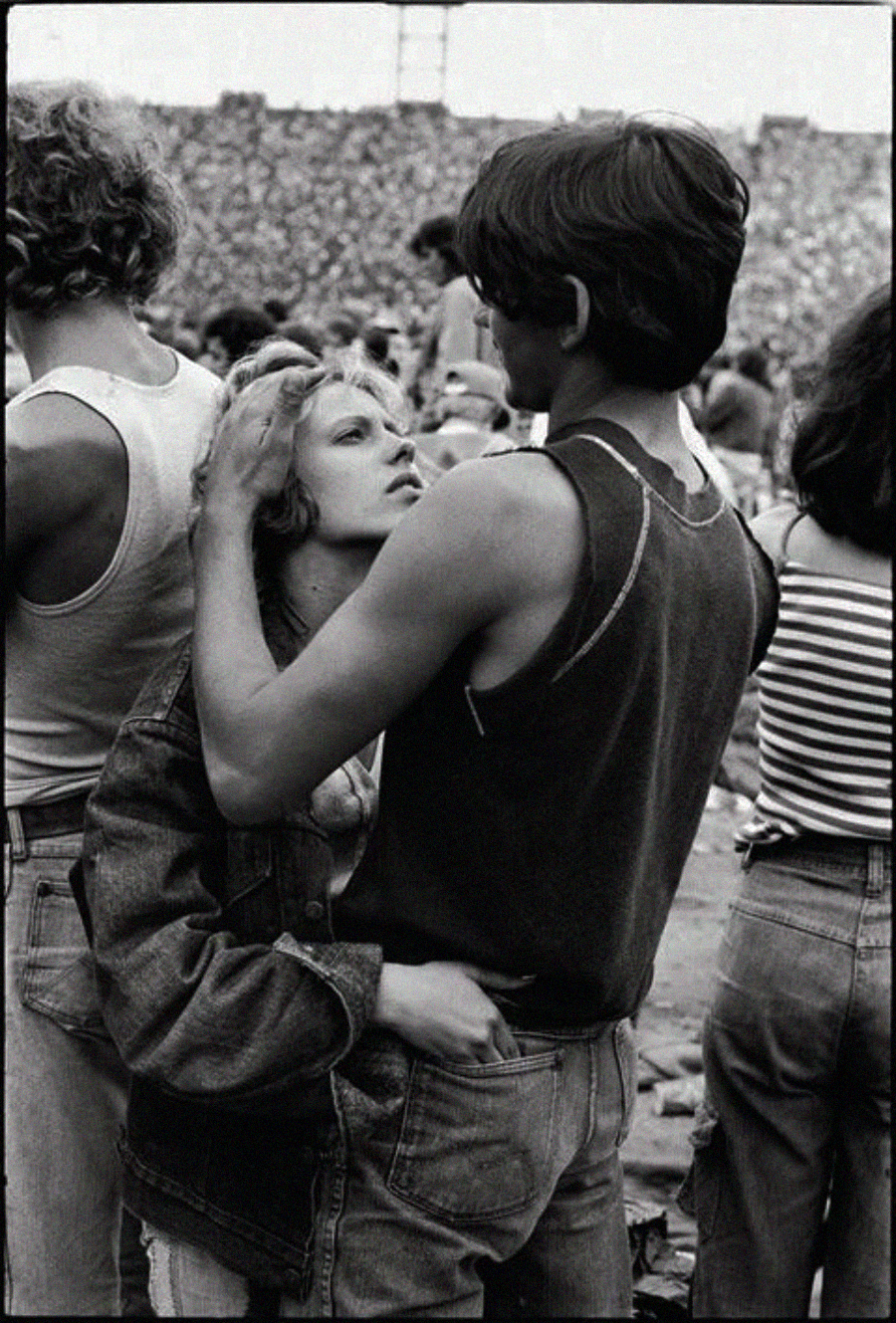 A young couple embraces closely in a crowded outdoor event. The woman looks up at the man as he gently holds her head, while people and a large audience fill the background. Black-and-white photograph.