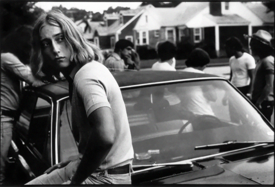 A young person with long hair sits on the hood of a car, looking to the side, while a group of people gather and talk in the background on a suburban street with houses. The photo is in black and white.