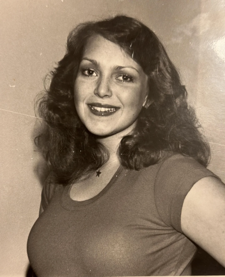 A young woman with wavy, shoulder-length hair smiles at the camera. She is wearing a short-sleeve shirt and a small necklace with a star pendant. The photo is in black and white.