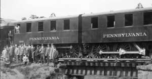 A group of men in suits and hats stand beside and in front of a Pennsylvania railroad train car on a bridge. The train car is dark-colored and labeled "PENNSYLVANIA" in large letters.