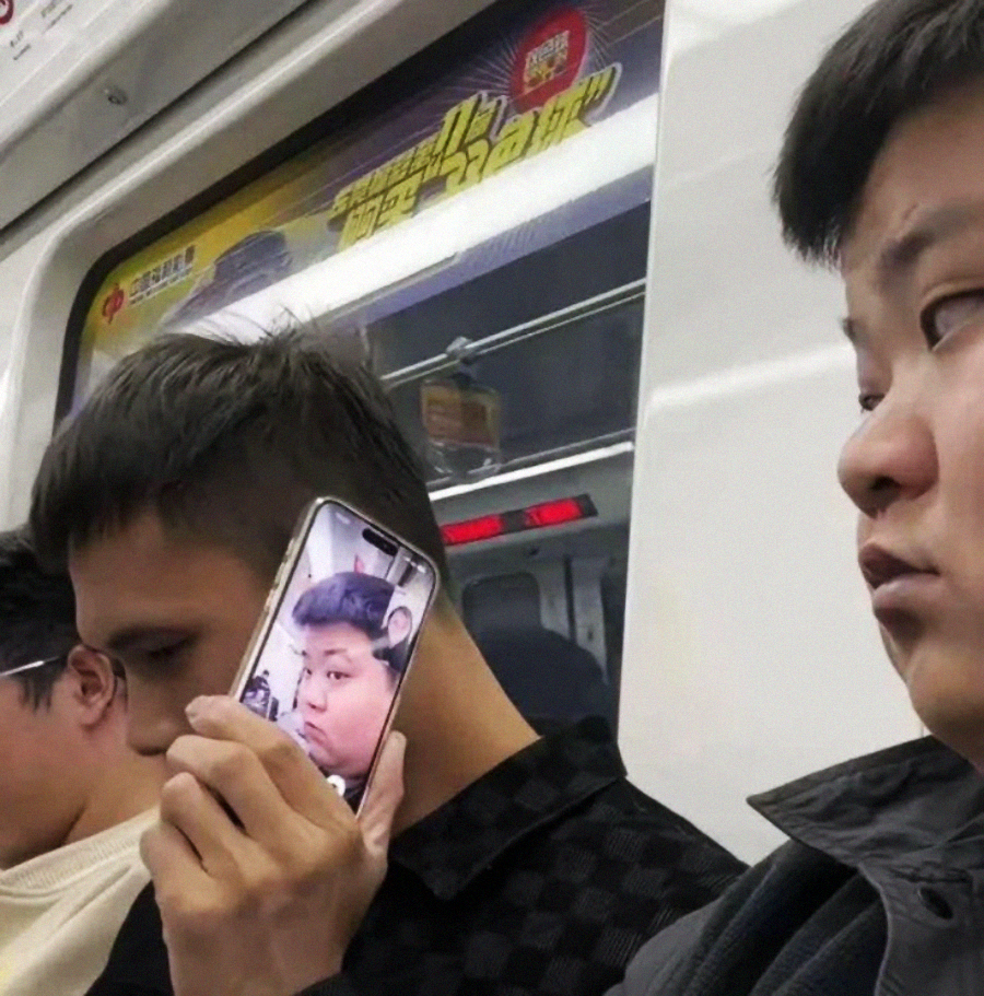 A person on a subway holds up a phone displaying another person’s surprised face, aligning it with their own head, while others sit nearby, unaware or uninterested.