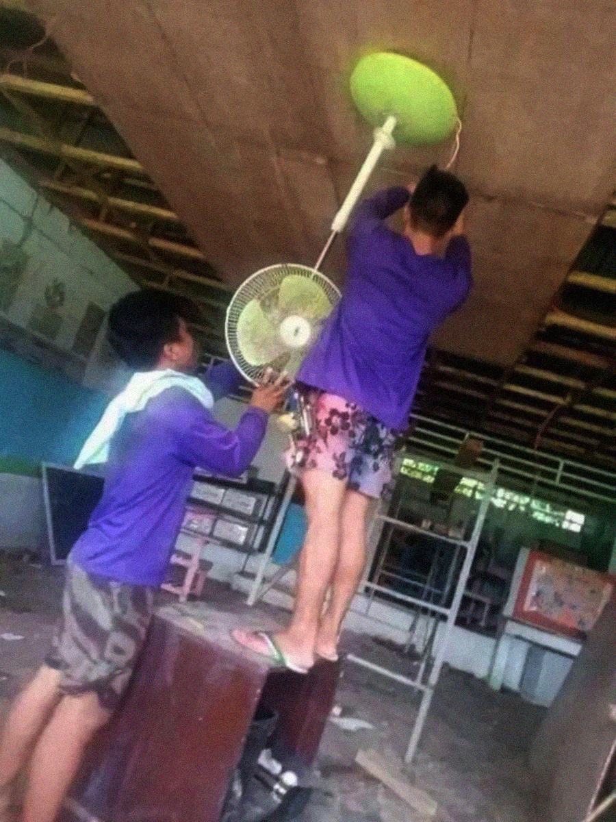 Two people in casual clothes and sandals are installing a ceiling fan; one stands on a desk while the other supports the fan. The room appears under construction or in disrepair.