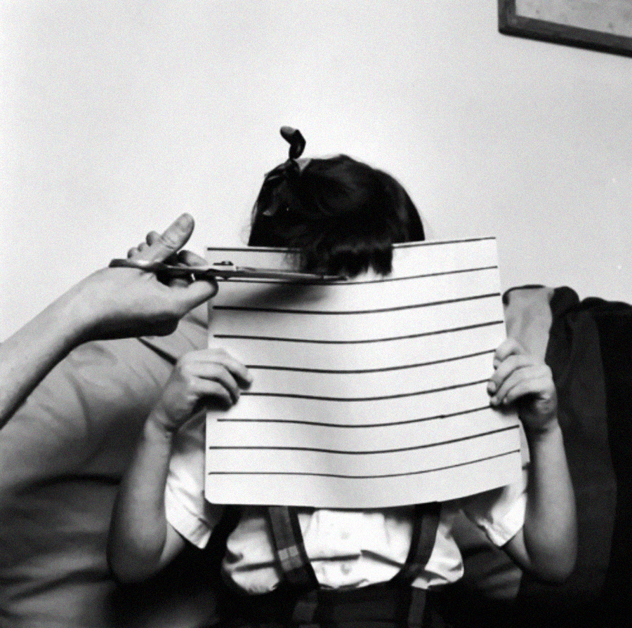 A child holds up a lined sheet of paper in front of their face while someone trims their bangs with scissors above the paper, using it as a cutting guide.