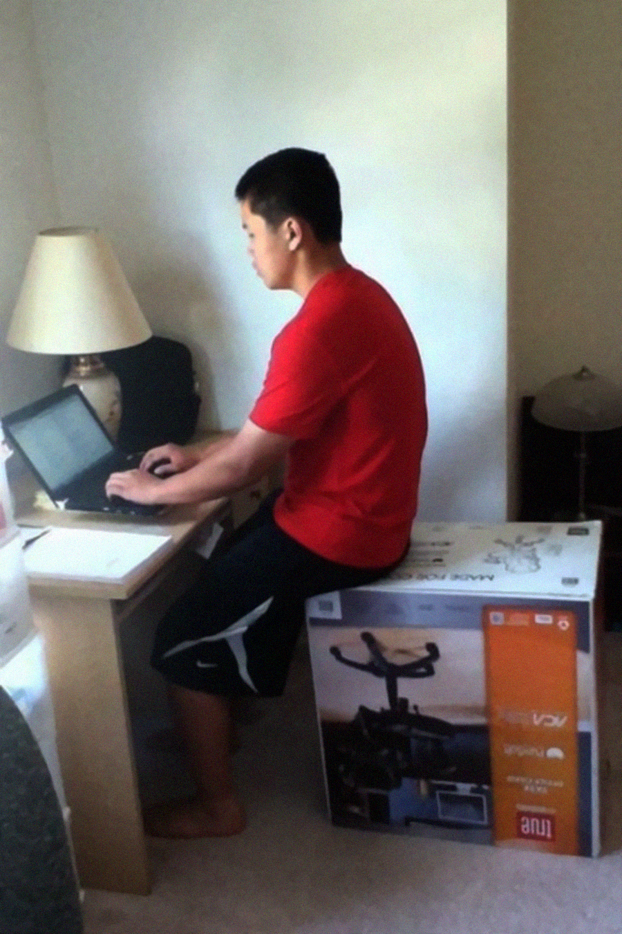 A person in a red shirt sits on a large exercise equipment box, using a laptop at a small desk in a corner of a room with a lamp, notepad, and another lamp on the floor nearby.
