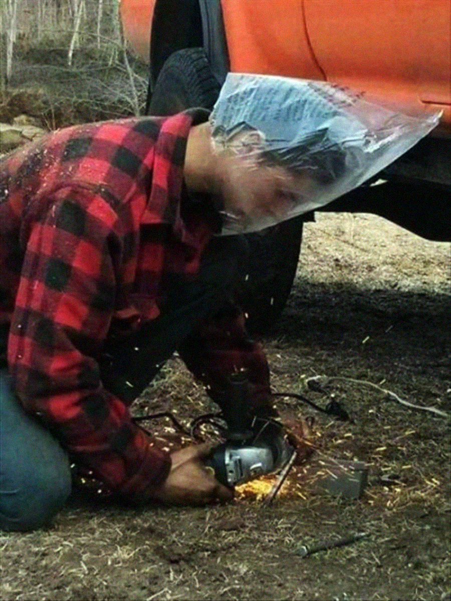 A person in a red plaid shirt uses a power tool under a vehicle, wearing a large plastic bag over their head as makeshift protection while sparks fly from the tool.