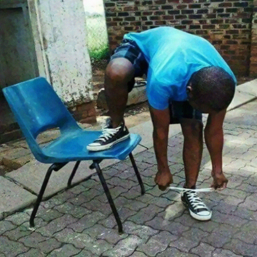 A person in a blue shirt and shorts ties their shoelace with one foot propped up on a blue plastic chair outdoors on a paved surface, next to a brick wall.
