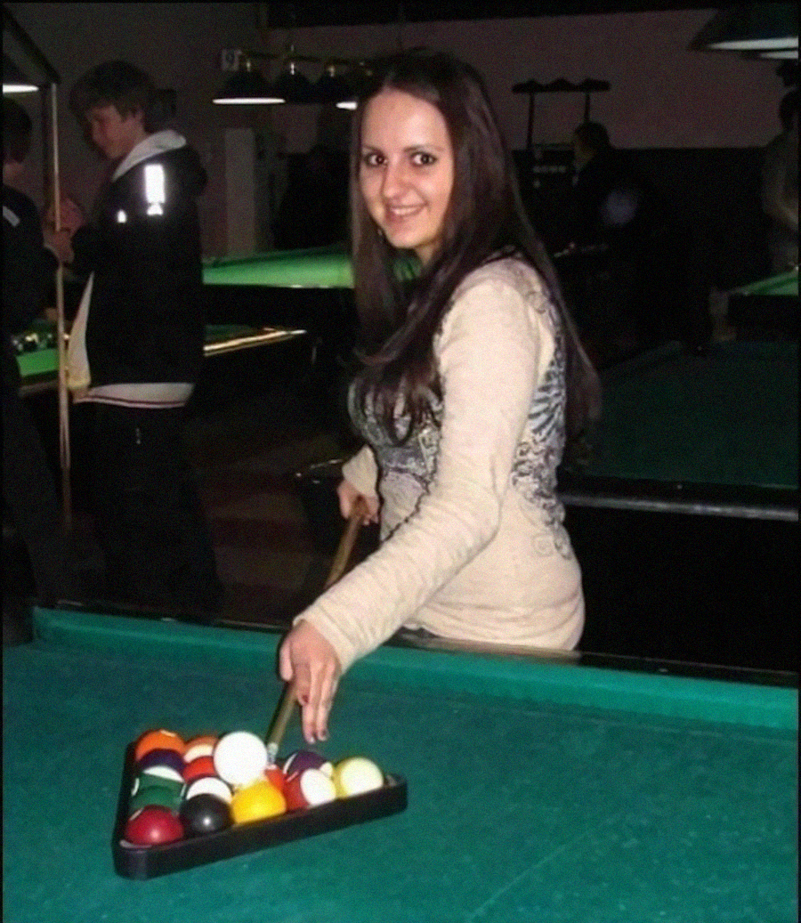 A woman with long brown hair smiles while setting up billiard balls in a triangle rack on a pool table. Other people and tables are visible in the dimly lit background.