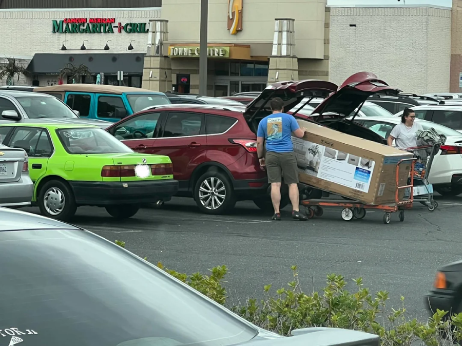 Two people in a parking lot struggle to fit a large boxed item from a shopping cart into the back of a red SUV, with other cars and store signs visible in the background.