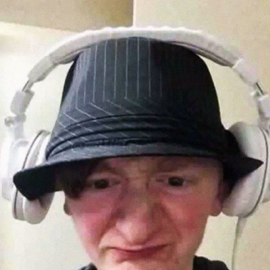 A person wearing a dark pinstripe fedora and large white headphones is making a scrunched facial expression, with a neutral indoor background.