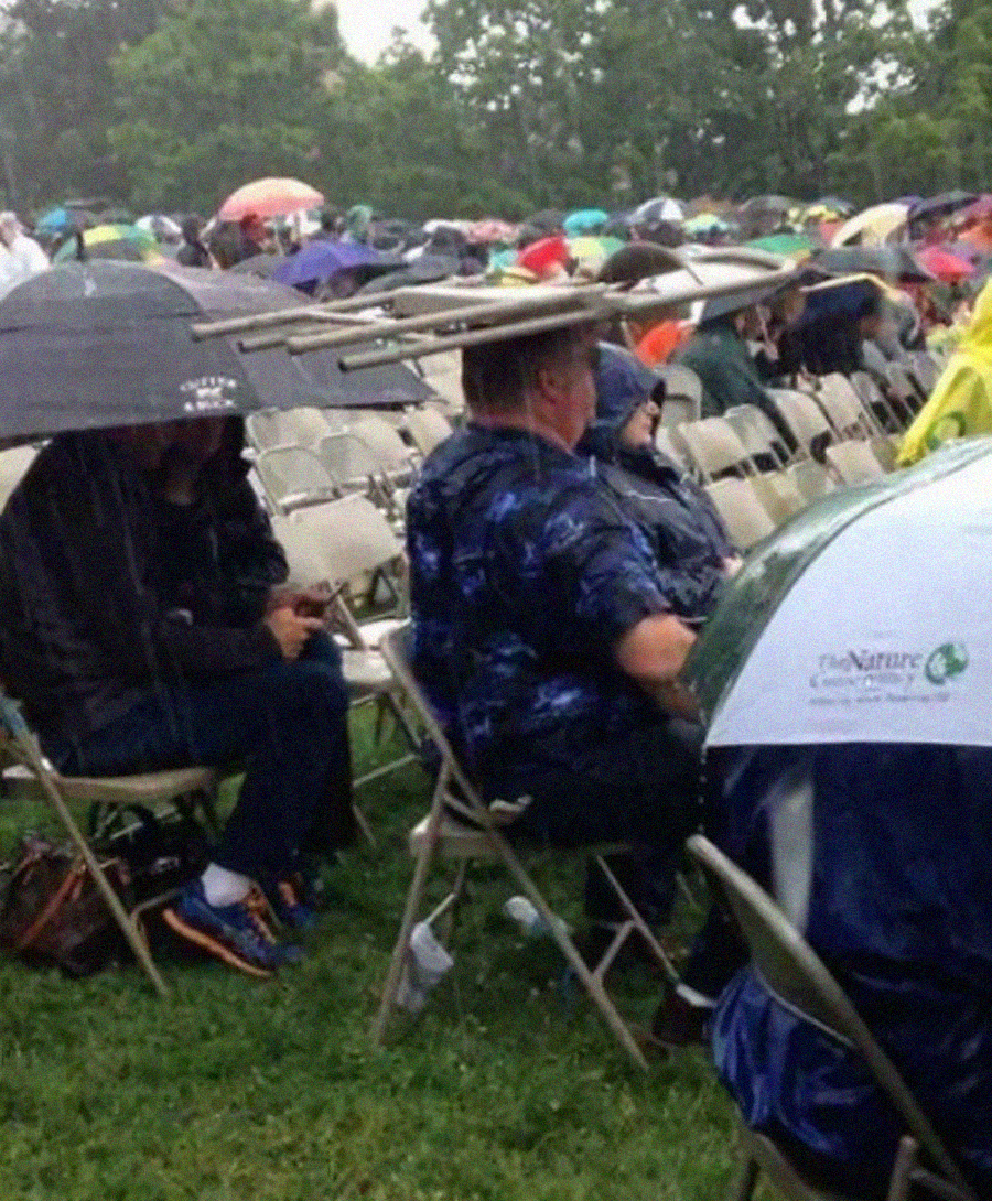 People sit outside in the rain at an event. Most use umbrellas, but one person has balanced two folding chairs on their head for cover. The crowd is seated on folding chairs, and trees are visible in the background.