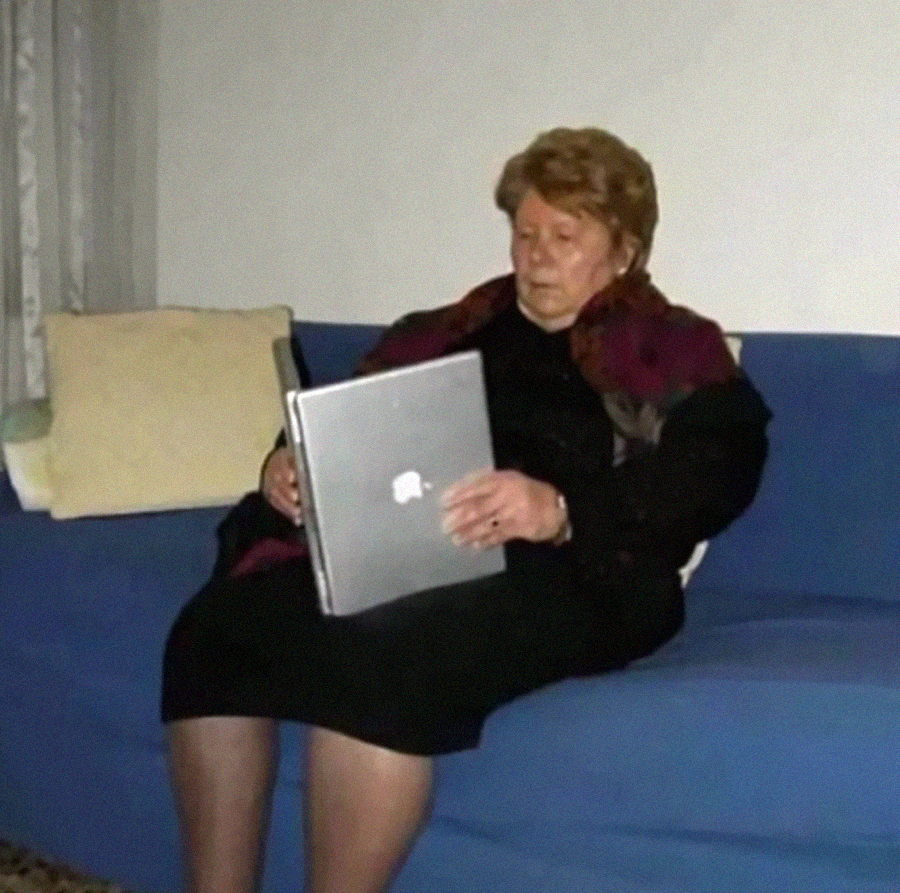 An elderly woman sits on a blue couch, holding a closed laptop in her lap and looking at the screen with a serious expression in a dimly lit room.