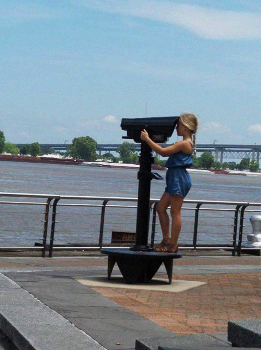 A woman in a blue romper stands on a raised platform looking through a public binocular viewer by a riverside, with water, trees, and a bridge visible in the background.