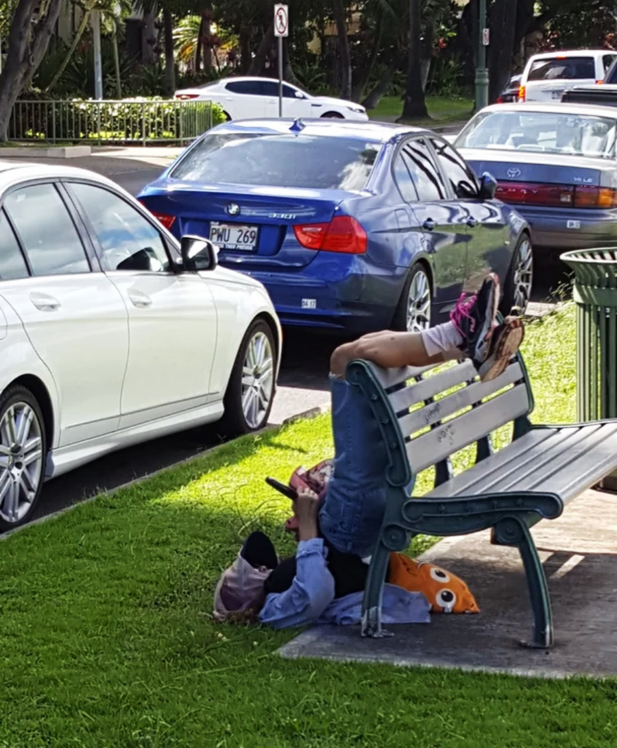 A person lies upside down on the grass with their legs resting on a park bench, holding a phone. Cars are parked nearby and an orange plush toy is next to them.