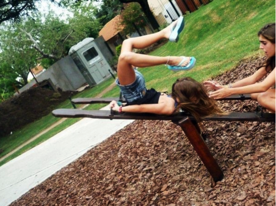 A girl in shorts and flip-flops is falling backward off a wooden seesaw, with her legs in the air, while another girl sitting on the ground nearby reaches out to catch her. They are in a park with grass and trees.