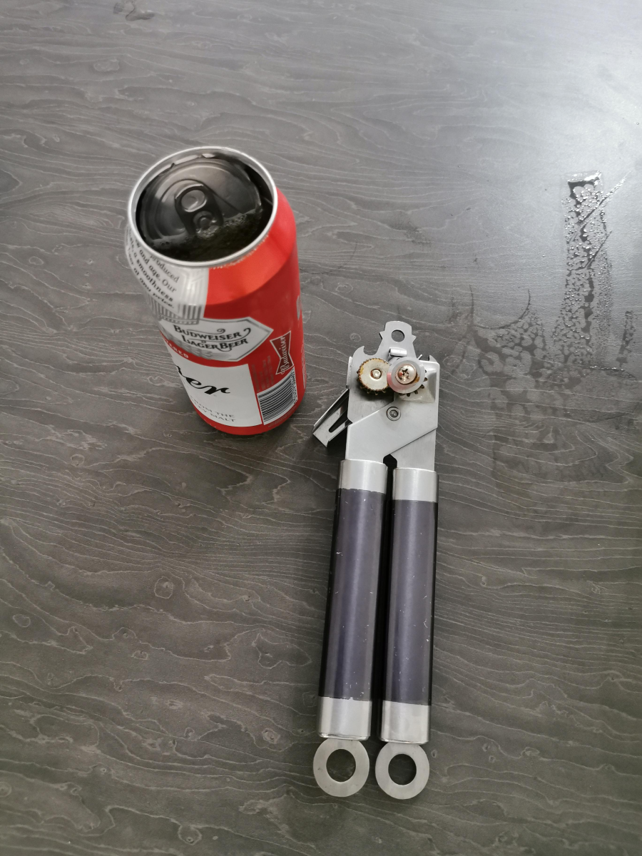 A red and white Budweiser beer can sits next to a silver and dark blue can opener on a dark wood grain surface.