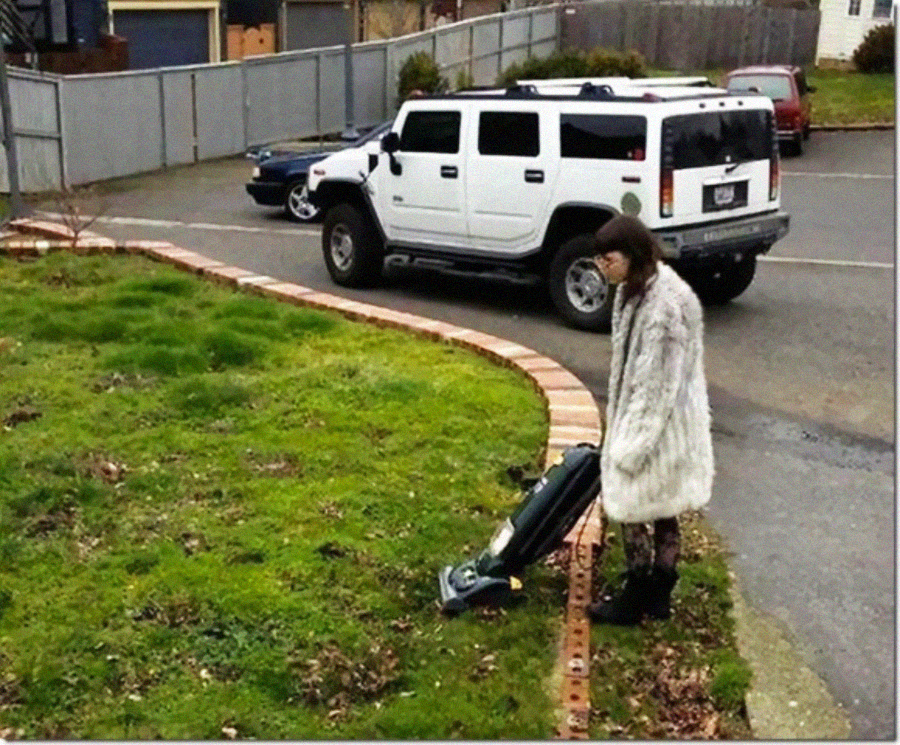 A person in a fur coat appears to be vacuuming a patch of grass in a yard, with a white SUV parked on the street behind them.