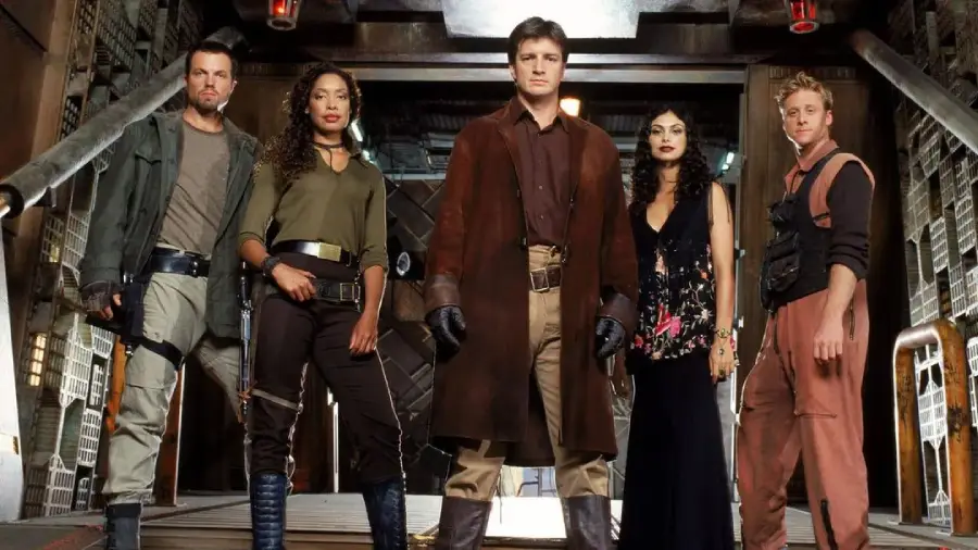 Five people in futuristic or sci-fi attire stand in a metal walkway, facing forward. The central figure wears a brown coat, while others wear a mix of utility gear and clothing, suggesting a science fiction setting.
