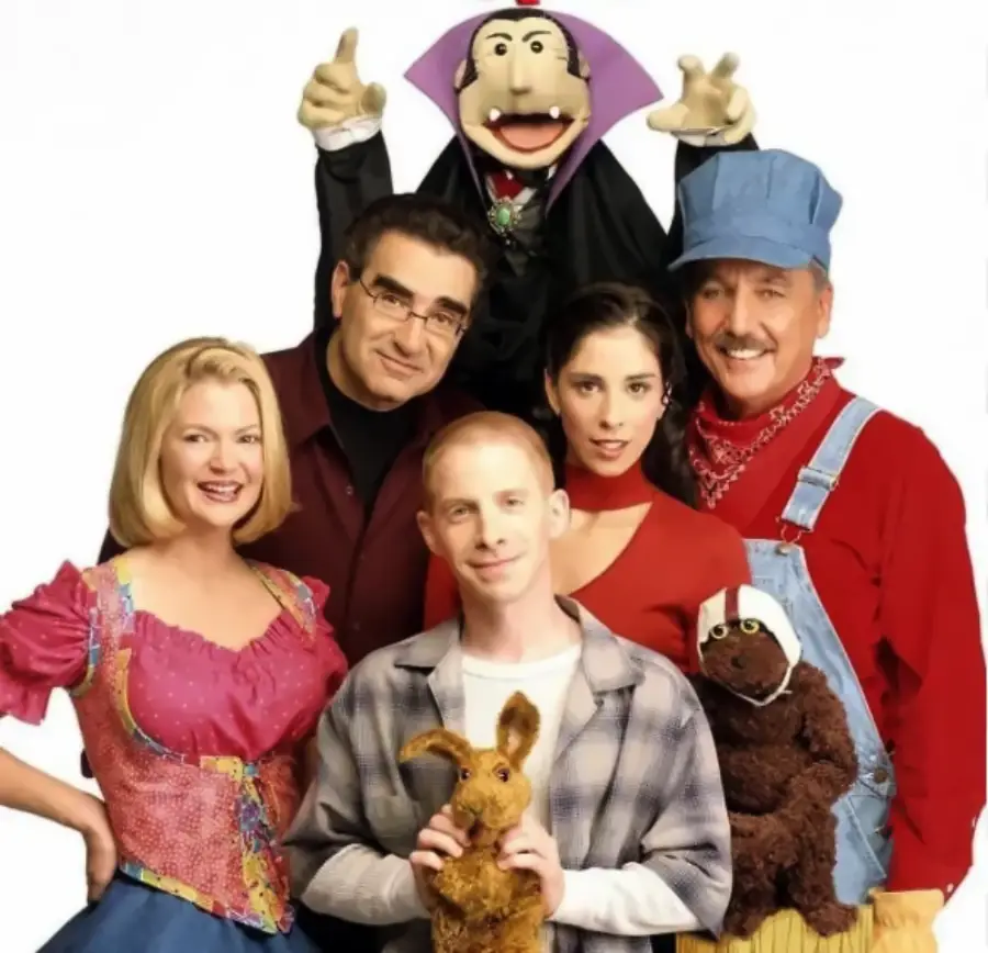 Five people pose together with two puppets: a man in a blue hat and overalls, a blonde woman in a pink top, a brunette woman in red, a bespectacled man in dark clothes, and a man in plaid holding a rabbit puppet.