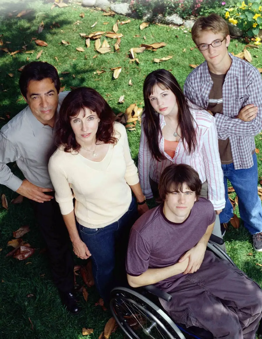 A group of five people, including a young man in a wheelchair, stand together outside on grass and look up at the camera; they appear to be a family scene.