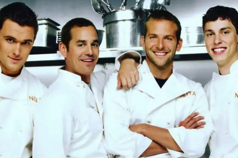 Four men wearing white chef jackets stand together in a kitchen, smiling at the camera with stainless steel pots and utensils in the background.