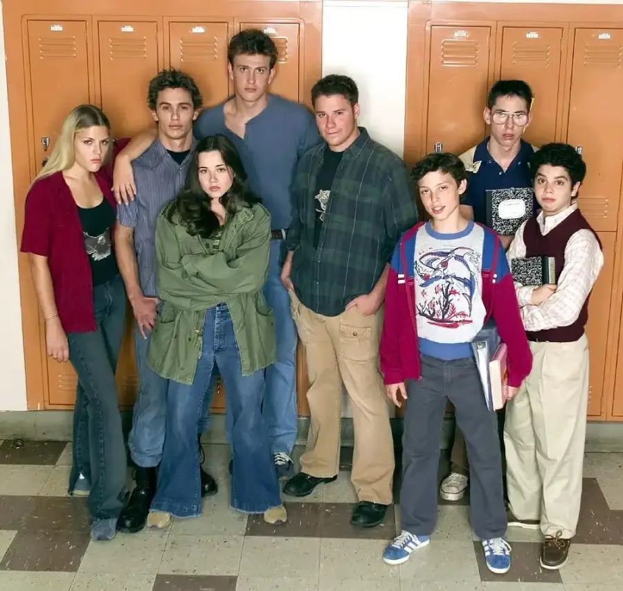 Eight teenagers stand in front of orange school lockers, dressed in casual 1980s-style clothing, some holding books, appearing as a high school group posing together in a hallway.