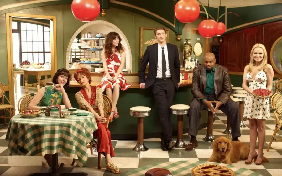 Six people, including three women sitting at a table, a man and a woman at a counter, and a man with a dog on the floor, pose in a retro diner decorated with giant cherries and several pies on display.