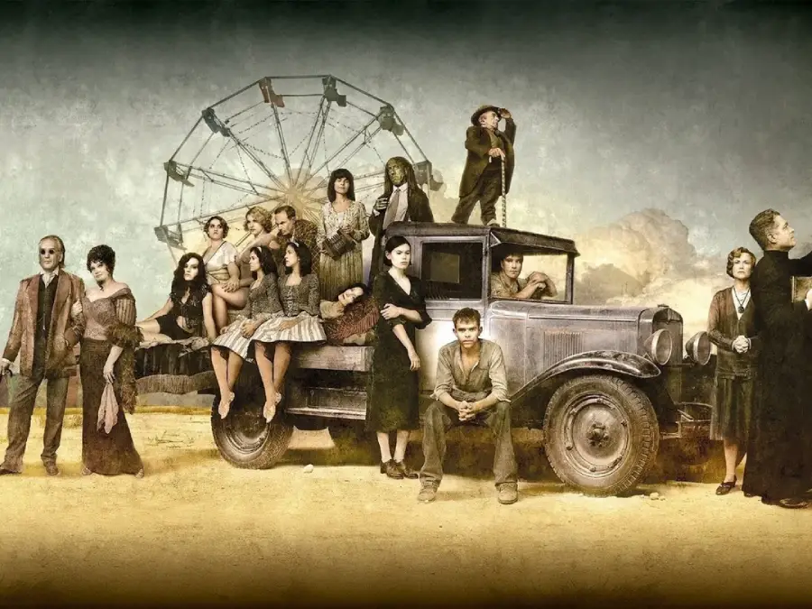 A group of people in vintage clothing pose around an old truck in a dusty, rural setting with a Ferris wheel in the background. Some sit on the truck while others stand, creating a somber, dramatic scene.