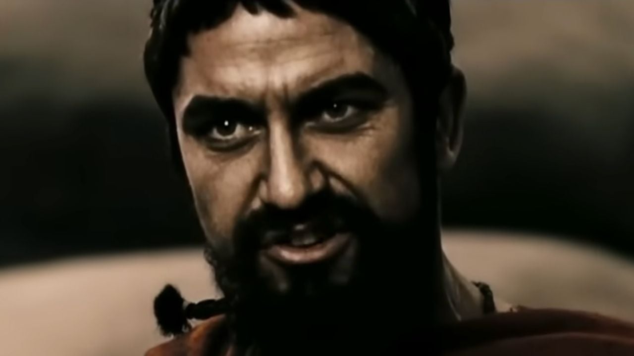 A man with a beard and intense expression looks forward. He has short, dark hair and wears a headband. The lighting is dramatic, casting shadows on his face.