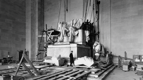 Workers assemble the Lincoln Memorial statue in a large, unfinished hall, using scaffolding, ropes, and wooden platforms. Marble pieces and construction materials are scattered around the base.