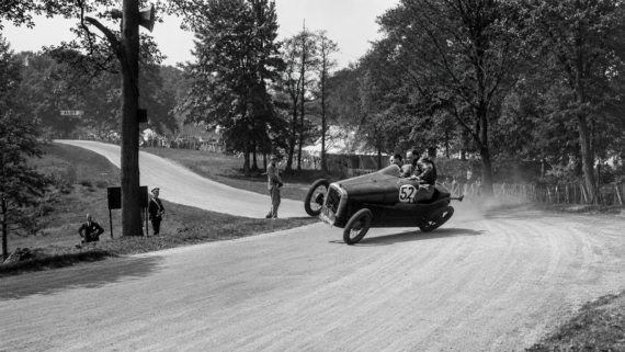 A vintage race car, number 52, turns sharply on a dirt road, lifting its left wheels off the ground. Three people are in the car, and trees line the road, with spectators visible in the background. 1920s