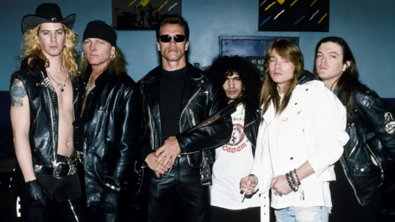 Six men pose together, mostly wearing black leather jackets and serious expressions. One man in sunglasses stands at the center holding a gun prop; another wears a white jacket. The background wall displays colorful patterns. 1990s