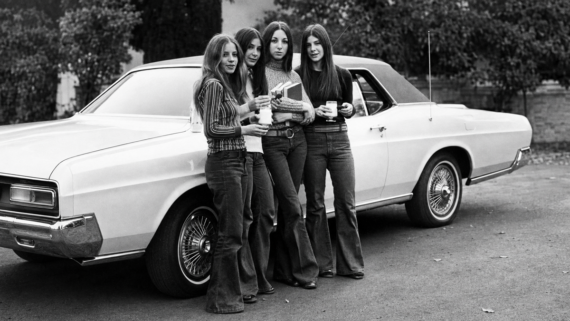 School. Four young women in bell-bottom jeans stand beside a classic car, holding drinks and fast food containers, smiling at the camera. The image is black and white and appears to be from the 1970s. 60s